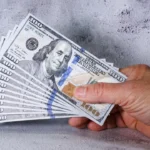 Hand holding fanned stack of $100 prop money bills demonstrating how to start investing with $100 in 2026