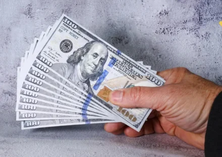 Hand holding fanned stack of $100 prop money bills demonstrating how to start investing with $100 in 2026